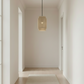 A modern hallway with light wood floors, a beige runner rug, and a woven cylindrical pendant light hanging from a black cord. The hallway features minimalist abstract wall art and a small wooden side table with a vase.