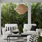 A modern tiered woven pendant light hangs in a bright, tropical-themed outdoor patio lounge area with white armchairs, a black coffee table, and lush green plants in the background.