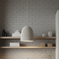 A woven pendant light with a textured, basketweave-like shade hangs from a rope cord in front of a gray brick wall with wooden floating shelves containing folded white towels and decorative jars.
