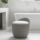 A round woven pouf stool with a white cushioned seat in a modern bathroom setting with a white freestanding bathtub and marble tiles.