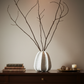 A white ribbed ceramic vase holding bare brown branches, displayed on a dark wooden console table against a warm beige wall, with stacked books and a small decorative tray nearby.