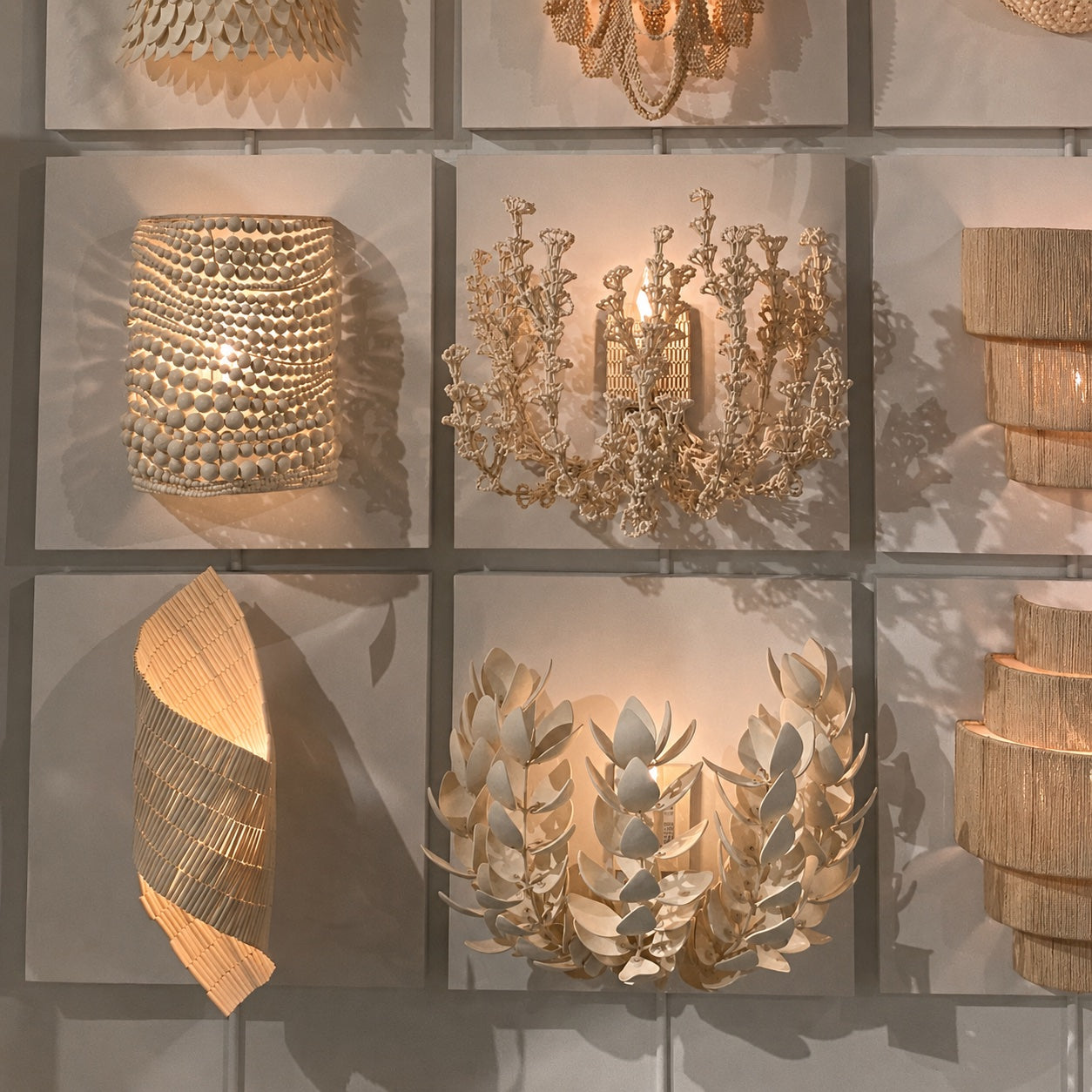 A collection of elegant, warm-toned decorative wall sconces and lamps displayed on a white tiled wall. The designs include a textured shell-like cylinder, a gold-toned crystal chandelier-style candle holder, a sculptural leafy wreath, a curved woven shell piece, and stacked woven shades, all casting a warm glow.