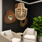 A woven rattan chandelier with three exposed light bulbs hangs in a modern living room corner with black walls, two beige armchairs, sunburst mirrors, and a large green plant.
