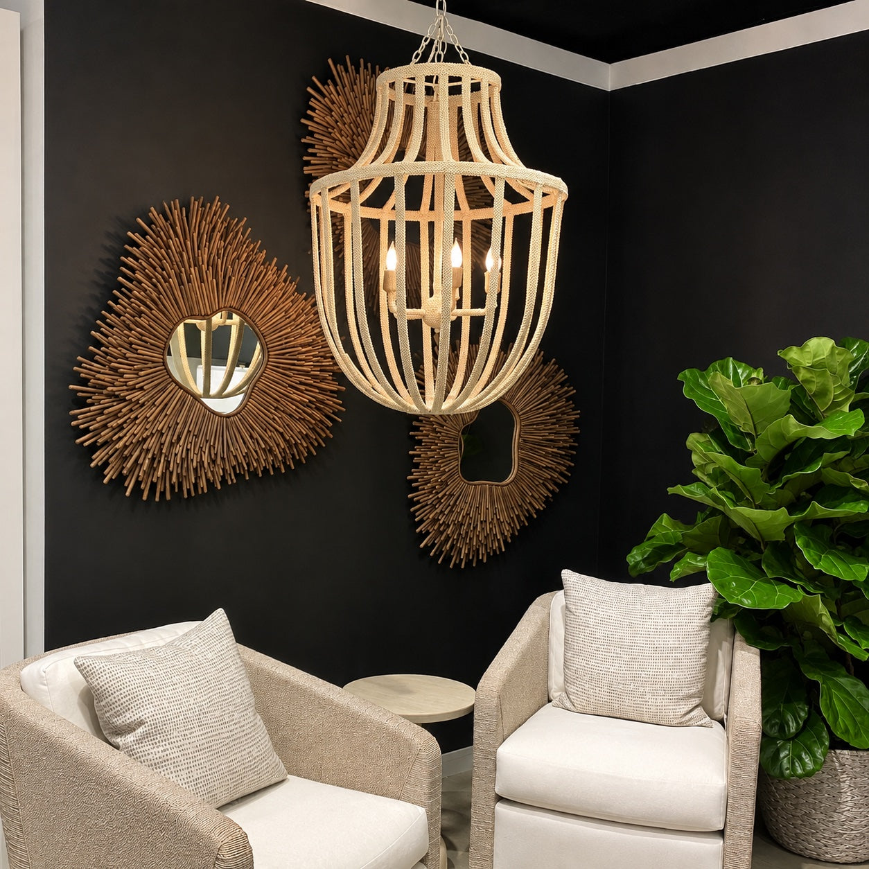 A woven rattan chandelier with three exposed light bulbs hangs in a modern living room corner with black walls, two beige armchairs, sunburst mirrors, and a large green plant.