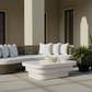 Outdoor patio furniture set featuring a curved wicker sofa with white cushions, a matching white coffee table, and several potted green plants against a beige building background.