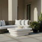 Outdoor patio furniture set featuring a curved wicker sofa with white cushions, a matching white coffee table, and several potted green plants against a beige building background.