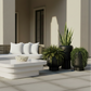Outdoor patio furniture set featuring a curved wicker sofa with white cushions, a matching white coffee table, and several potted green plants against a beige building background.