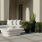 Outdoor patio furniture set featuring a curved wicker sofa with white cushions, a matching white coffee table, and several potted green plants against a beige building background.