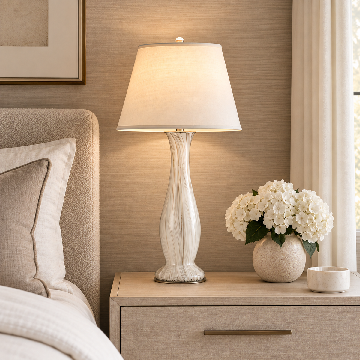 A lit table lamp with a textured white ceramic base and a warm cream fabric shade sits on a light wood nightstand in a bedroom setting, next to a bed and a vase of white flowers.
