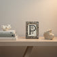 A rectangular picture frame with a textured, speckled stone-like border in beige, brown, and gray tones, featuring a large black decorative letter 'P' on an off-white insert.