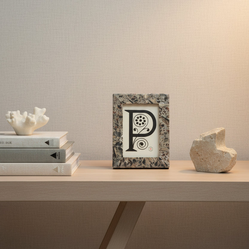 A rectangular picture frame with a textured, speckled stone-like border in beige, brown, and gray tones, featuring a large black decorative letter 'P' on an off-white insert.