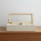 A light natural wood watch box with a clear glass lid, gold-tone hinges, and a cream-colored interior cushion holding five watch slots, shown open on a light gray background.