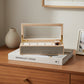 A luxury watch display box with a light wood frame, a clear glass lid, gold-tone hinges, and a textured gray shagreen front panel. Inside, the box features a cream-colored velvet lining with individual slots and a single gold watch with a dark brown leather strap is displayed on the center cushion.
