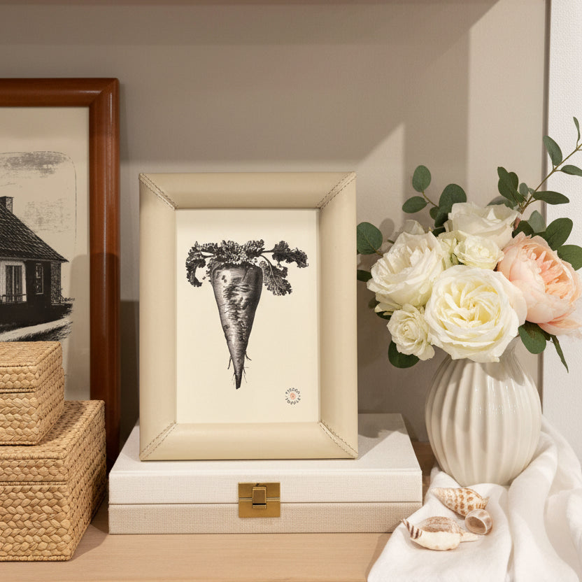 Two framed art prints displayed on a shelf. The front frame is cream-colored faux leather and features a black-and-white illustration of a carrot with leafy greens.