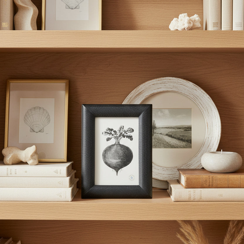 A black textured leather picture frame containing a black and white botanical illustration of a turnip, on a bookshelf with other frames and decor.