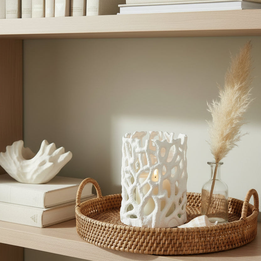A white textured candle holder with an organic, coral-like openwork pattern, containing a cream-colored candle on a bookshelf