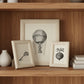 Three small framed art prints with light wood frames on a wood background. The center print is larger and shows a vintage-style hot air balloon illustration.