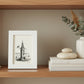 A small white textured picture frame holding a black ink illustration of a lighthouse over water, photographed against a bookshelf