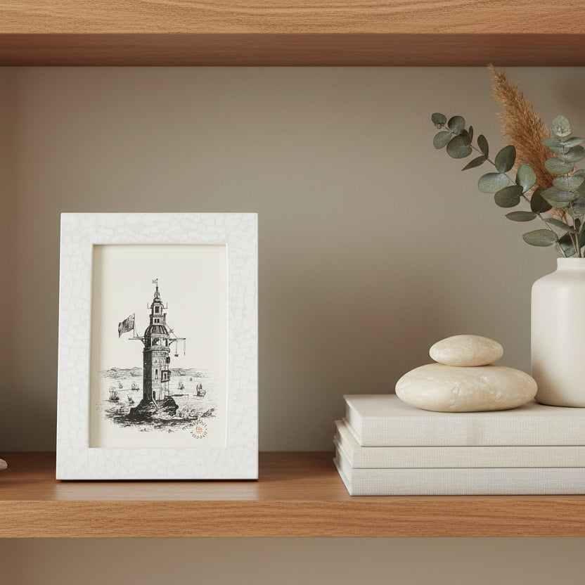A small white textured picture frame holding a black ink illustration of a lighthouse over water, photographed against a bookshelf