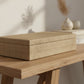 A rectangular storage box with a textured linen or burlap-style cover, shown closed against a white wall on a wood console table.