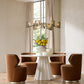 Modern dining room with a white round table and brown chairs, featuring a chandelier and floral arrangement.