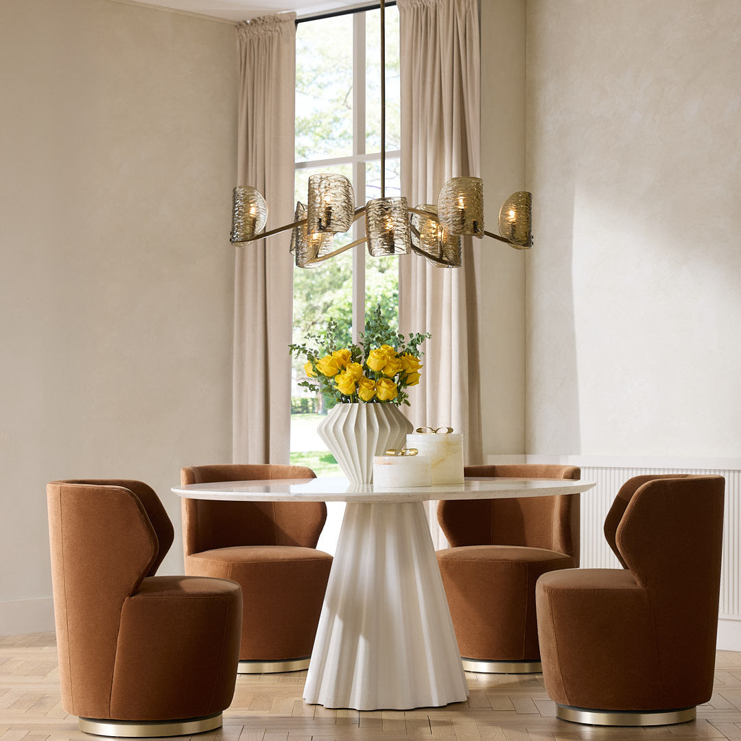 Modern dining room with a white round table and brown chairs, featuring a chandelier and floral arrangement.