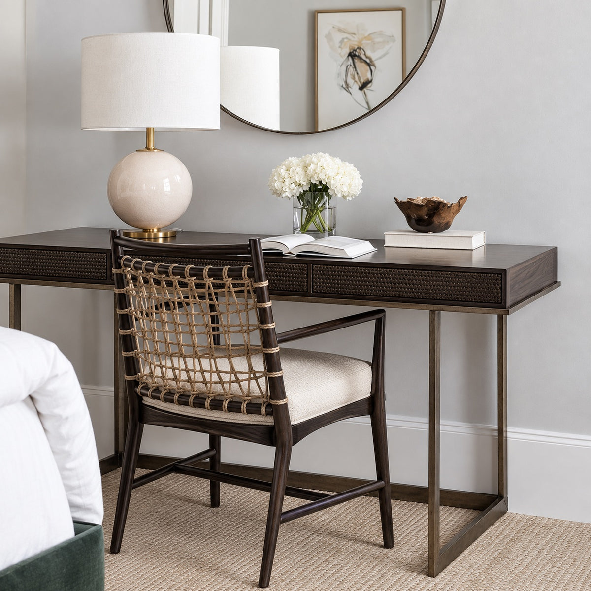 A dark wood writing desk with a woven-back chair, styled in a bedroom setting with a lamp, books, and a vase of white flowers.