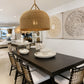 A large woven dome pendant light hangs over a dark wood dining table with woven-back chairs in a bright, modern dining room. The room features white walls with large circular woven wall art and a visible showroom in the background.