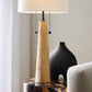 Beige cone-shaped lamp on a table with a black vase and white chair in the background