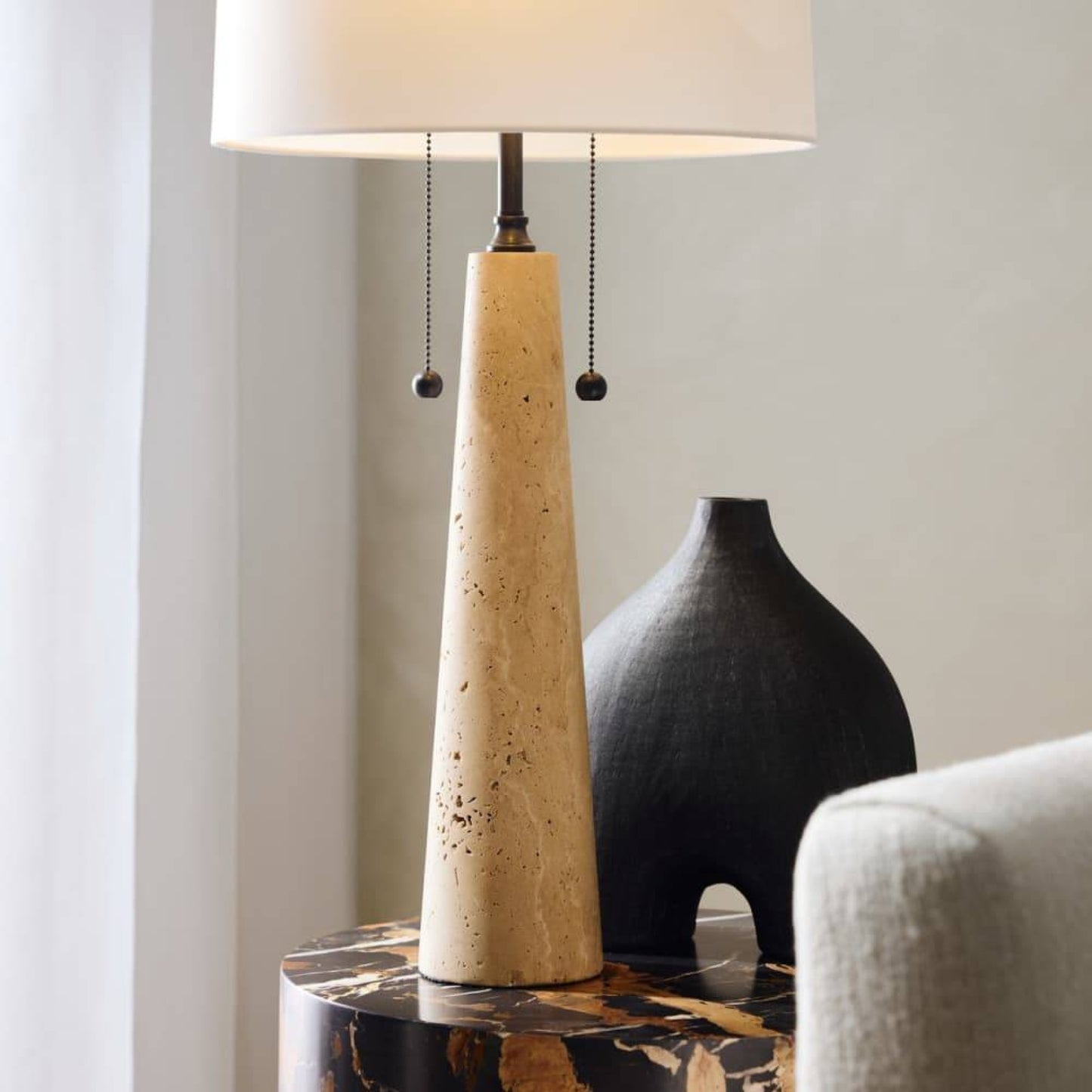 Beige cone-shaped lamp on a table with a black vase and white chair in the background