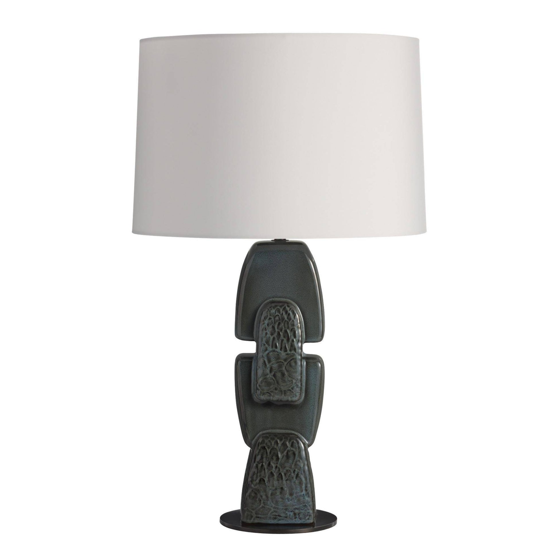 A table lamp with a large white drum shade and a sculptural ceramic base in a dark charcoal gray with subtle blue-green glaze accents, set against a plain white background.