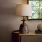 Table lamp with textured base and beige shade on a wooden surface near a window.