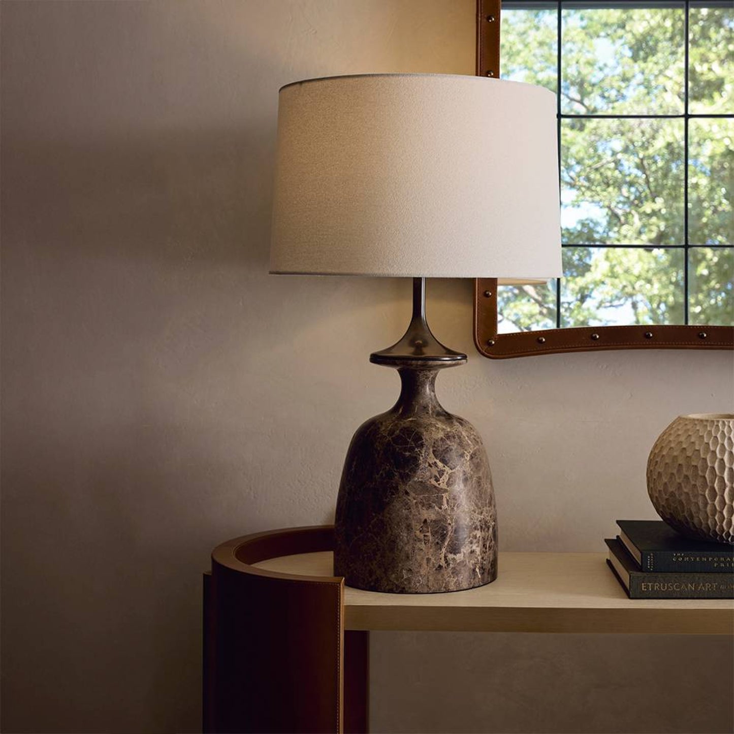 Table lamp with textured base and beige shade on a wooden surface near a window.