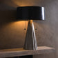 Table lamp with black shade and metallic base on a wooden surface against a textured wall.