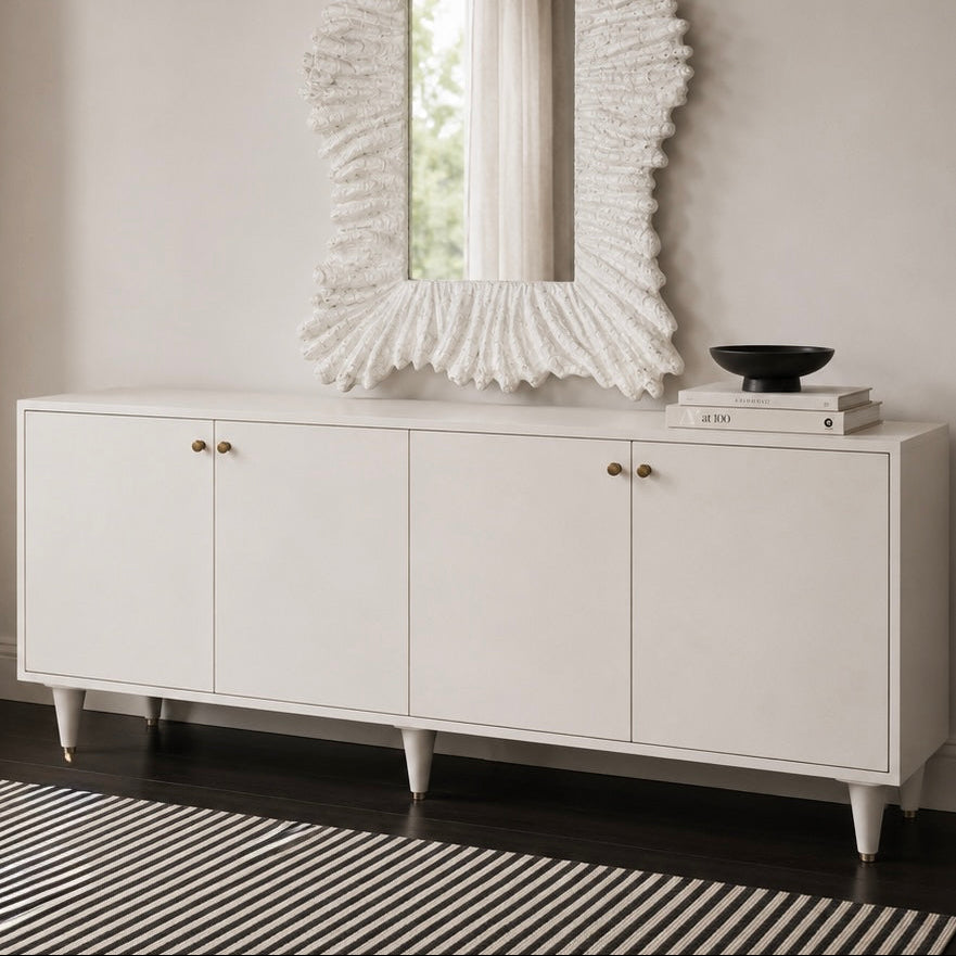 A white mid-century modern sideboard cabinet with four doors and small brass knobs, styled with a matching white textured sunburst mirror on top and a black bowl on books. The cabinet stands on dark wood flooring over a black and white striped rug against a neutral beige wall.