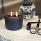 Lit candle in a black container with 'Ratner & Rose' branding on a textured surface.