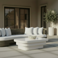 Modern outdoor patio furniture set featuring a curved wicker sofa with white cushions, a low white rectangular coffee table, and a decorative bowl, set against a neutral beige wall with large window panes and a potted olive tree.