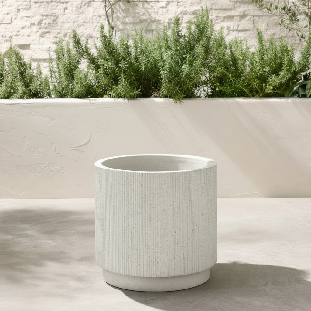 A cylindrical white textured planter pot with a ribbed exterior, set on a light concrete patio in front of a white brick wall and green plants.