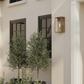 A modern outdoor wall sconce light fixture with a warm brass finish and a lit glass lantern-style shade, mounted on a beige stucco house exterior next to dark-framed windows and green plants.