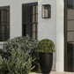 A black outdoor wall sconce light fixture mounted on a white brick house exterior, positioned above a large black planter with a green topiary and next to a white column and dark-framed windows.