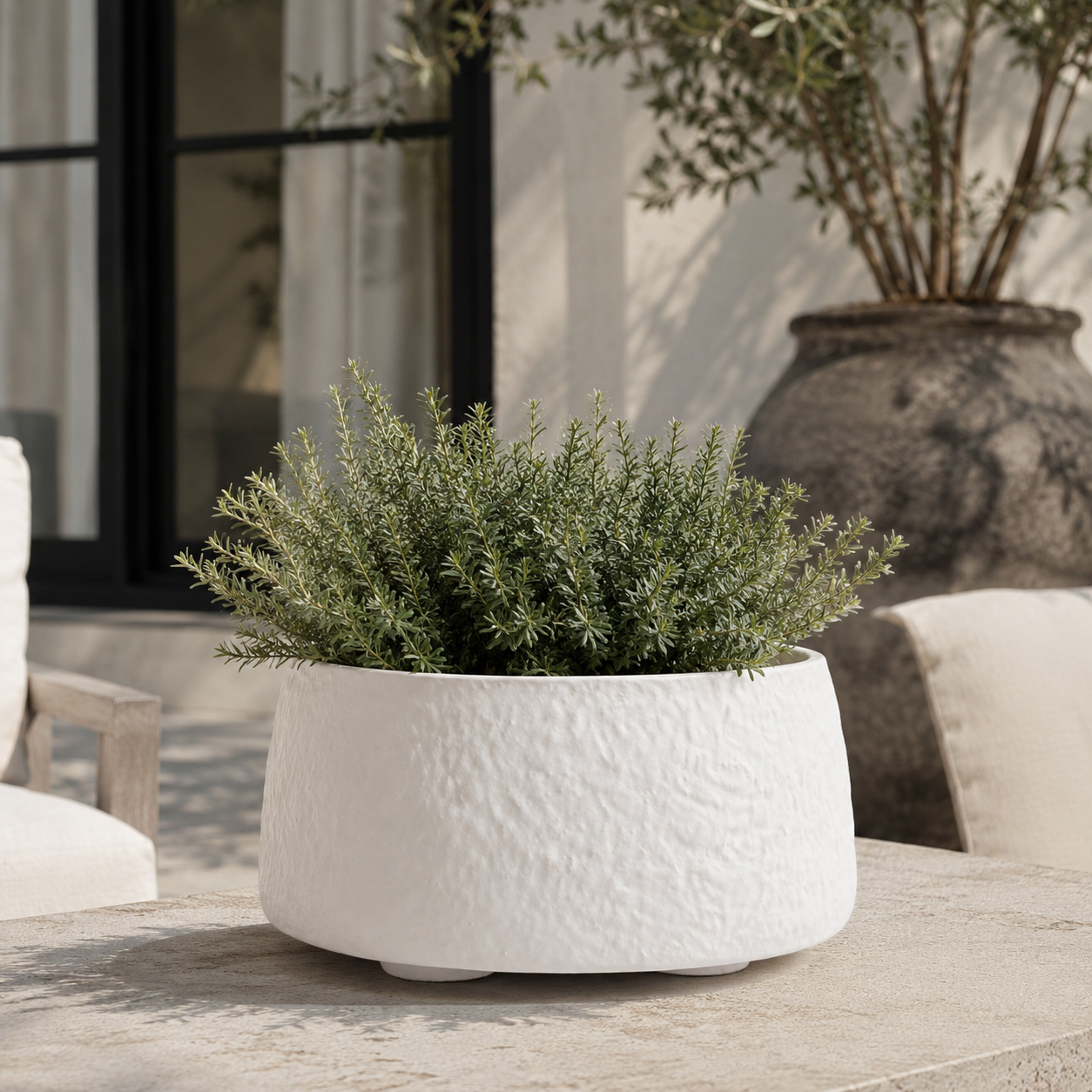 A textured white ceramic planter with a green leafy herb plant, displayed on a stone patio table in an outdoor patio setting.