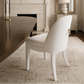 A white upholstered dining chair with a curved back and gold-toned ring detail, positioned in a classic dining room setting with a fireplace, mirror, and table.