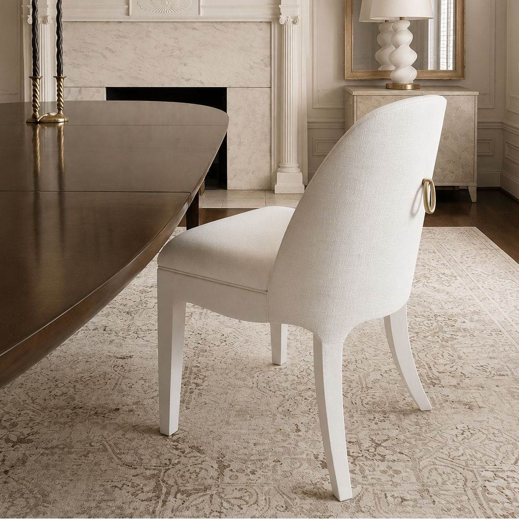 A white upholstered dining chair with a curved back and gold-toned ring detail, positioned in a classic dining room setting with a fireplace, mirror, and table.