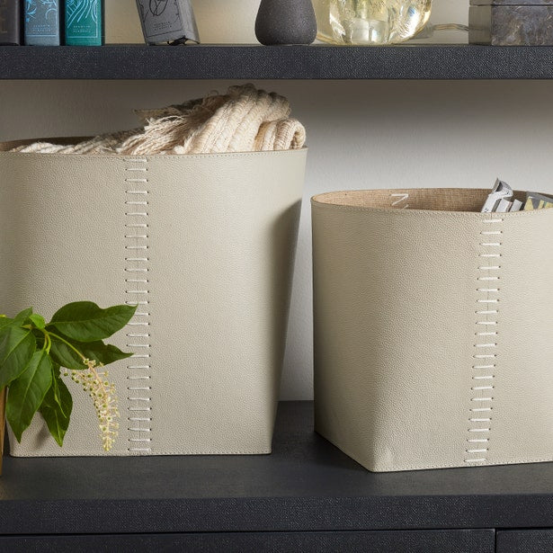 Two beige storage baskets on a dark surface with a plant and books in the background.