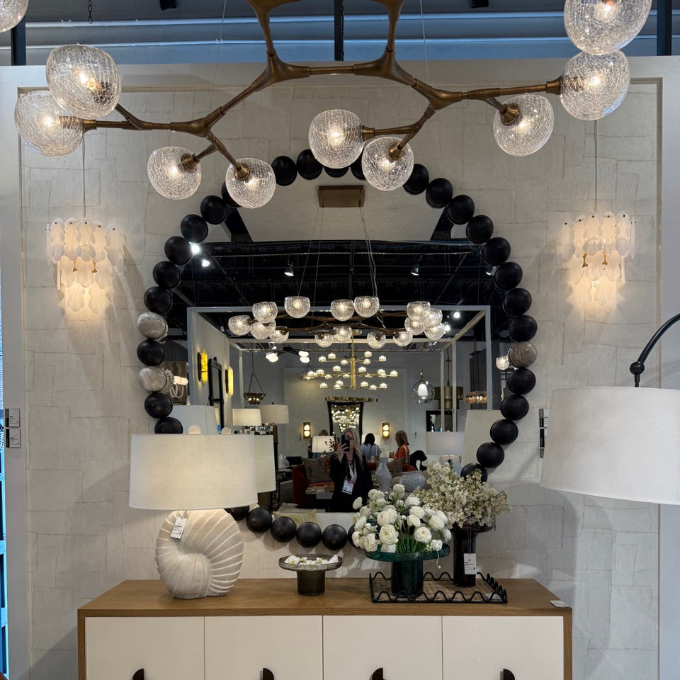 A large round mirror with a black beaded frame hangs above a white and wood console cabinet in a styled interior. The mirror reflects a showroom ceiling with multiple hanging lights, and the cabinet is decorated with a shell-shaped lamp, floral arrangements, and decorative objects.