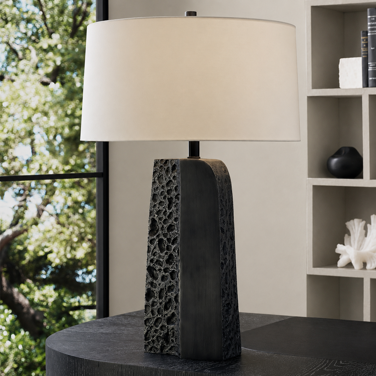 A modern table lamp with a textured, porous dark charcoal base and a large cream-colored drum shade, set on a dark round table in a styled interior room.