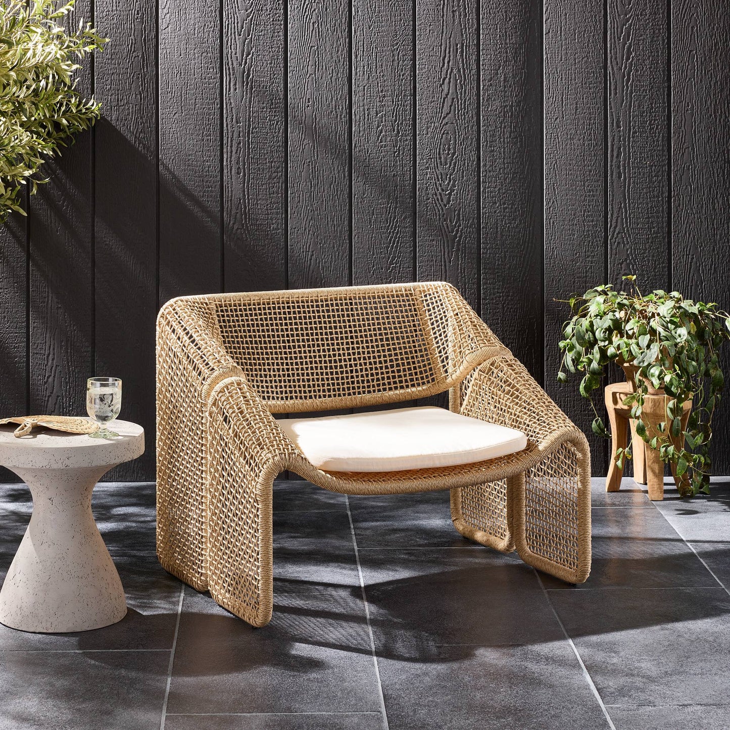 Woven chair with a cushion on a patio with a dark wooden wall and plants.