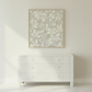 A square framed wall art print with a light gray and white leaf pattern, displayed above a white three-drawer dresser against a white wall on a light herringbone floor.