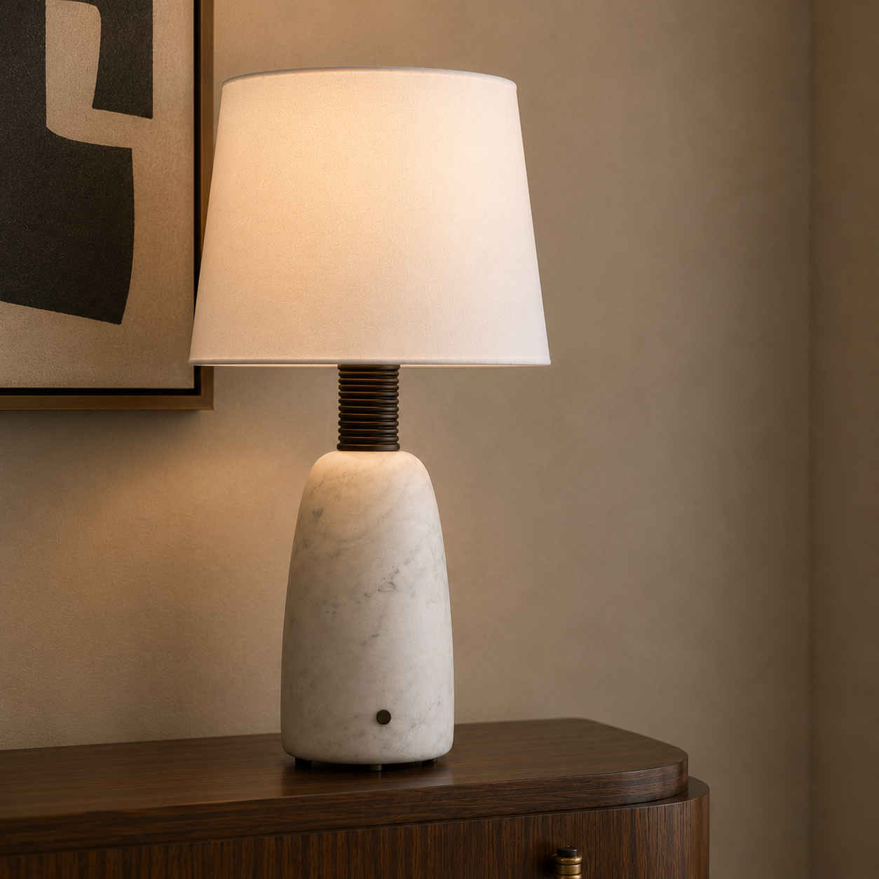 A table lamp with a white marble base and a white fabric drum shade, illuminated with a warm glow. The lamp is styled on a dark wood sideboard against a beige wall with a framed print partially visible.