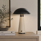 A mushroom-shaped table lamp with a matte black metal dome shade and a textured beige stone-like base, styled on a light wood sideboard next to a vase and books.